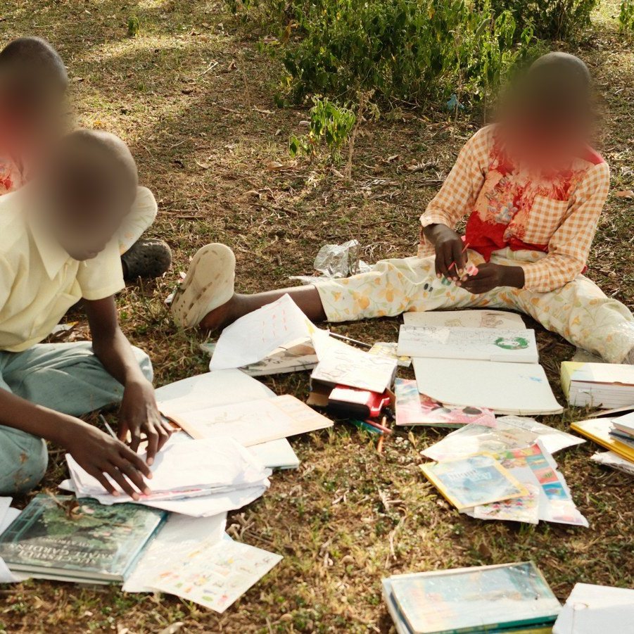 An outdoor art session in Kenya being filmed, children working with books and drawings, faces blurred for privacy